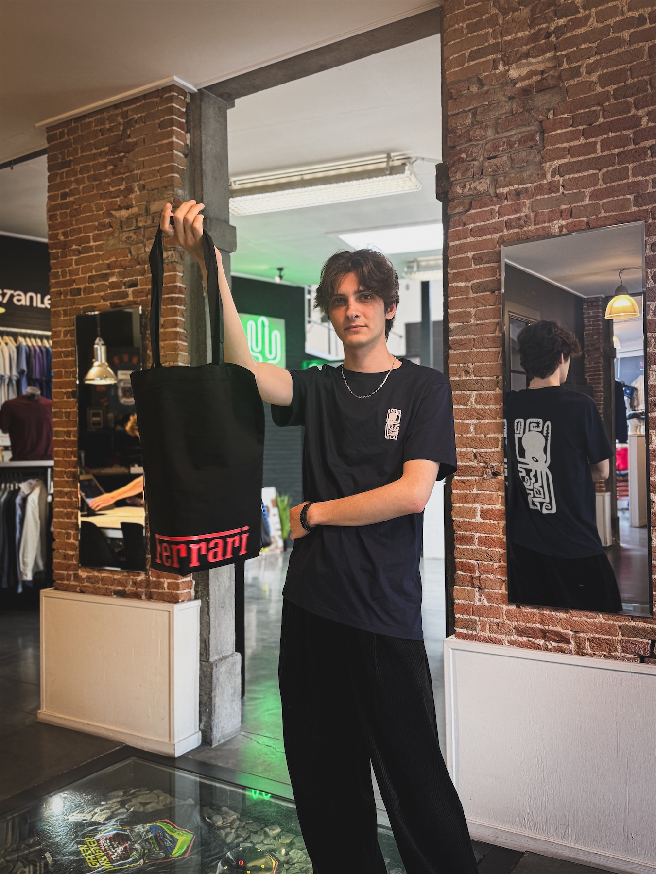 Andrei stands between two brick pillars inside the PAS PRINT store, holding a black tote bag with red Ferrari lettering. His dark blue T-shirt features a small white graphic on the chest and a larger version on the back, visible in the mirror.